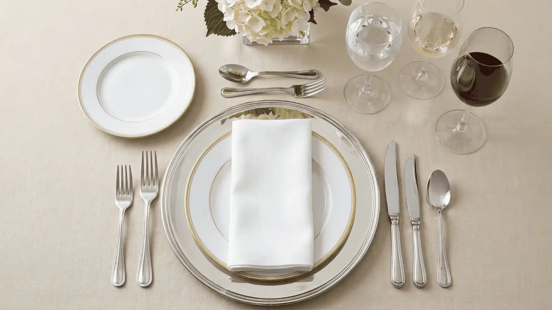 A formal table setting with charger plate, dinner plate, flatware, dessert utensils, glassware, and napkin in a refined, elegant layout.