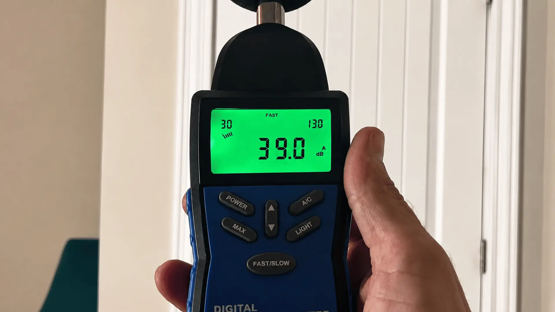 Hand holding a digital sound level meter showing 39.0 dB in front of a white interior door with soft indoor lighting near door