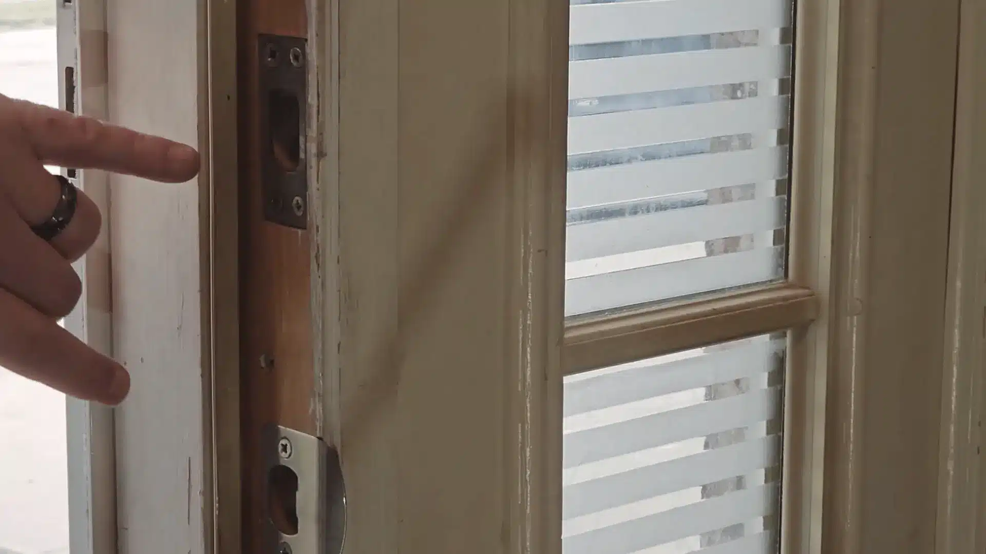 Finger pointing at door strike plates on a wooden frame while aligning hardware near a glass window with blinds.
