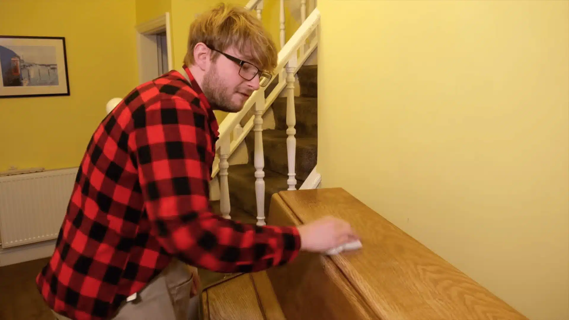 A man wearing glasses and a red checkered shirt polishing a wooden staircase banister with a cloth in a warmly lit hallway.