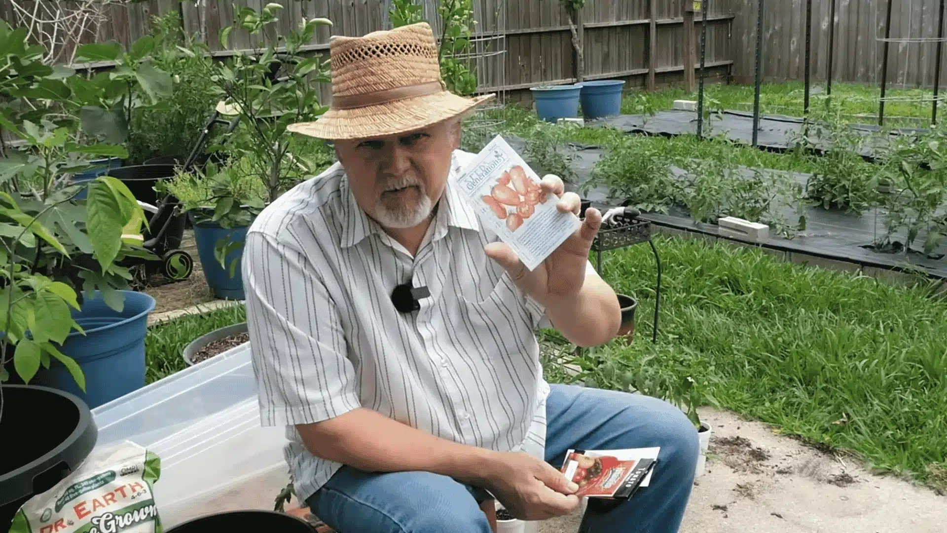 Man holding tomato seed packets while sitting in a backyard vegetable garden with raised beds and potted plants