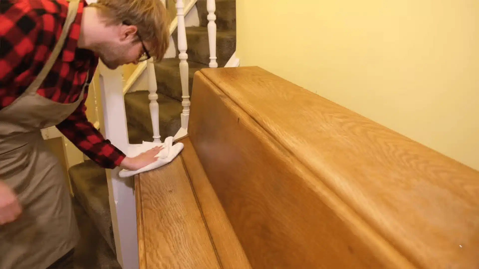 A person in a red checkered shirt polishing a wooden staircase banister with a white cloth, focusing on cleaning and restoring the wood surface.