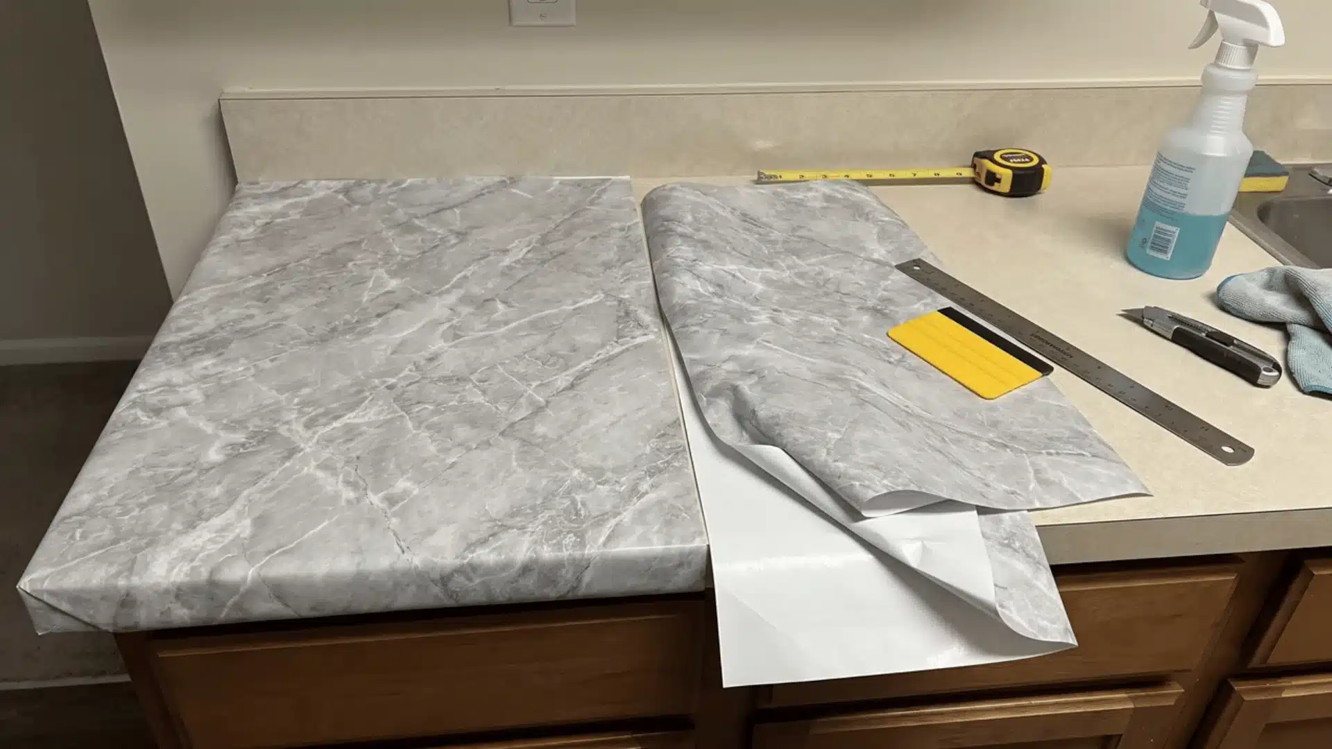 Contact paper partly applied to a countertop with backing exposed and a squeegee, knife, and straightedge nearby