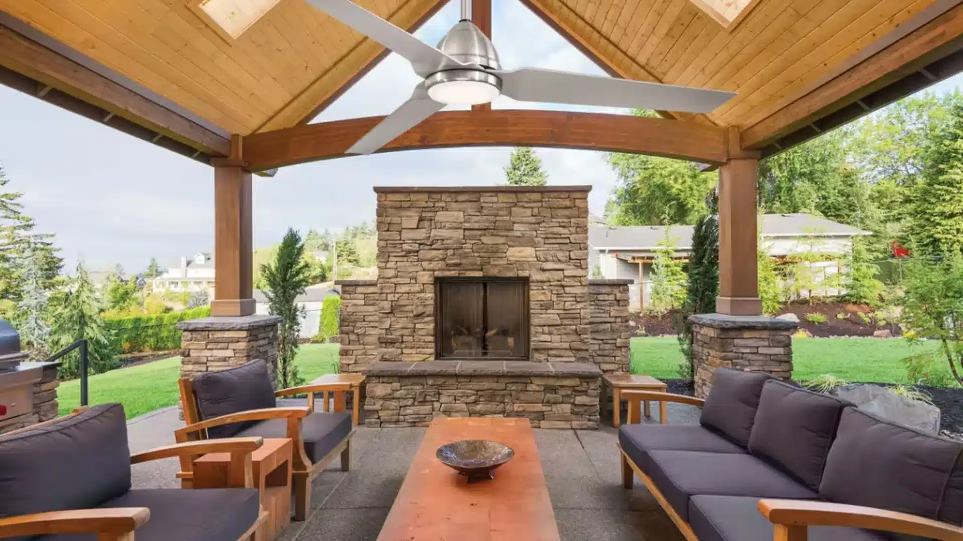 Covered outdoor patio featuring high wooden vaulted ceiling, central ceiling fan, and stone fireplace.