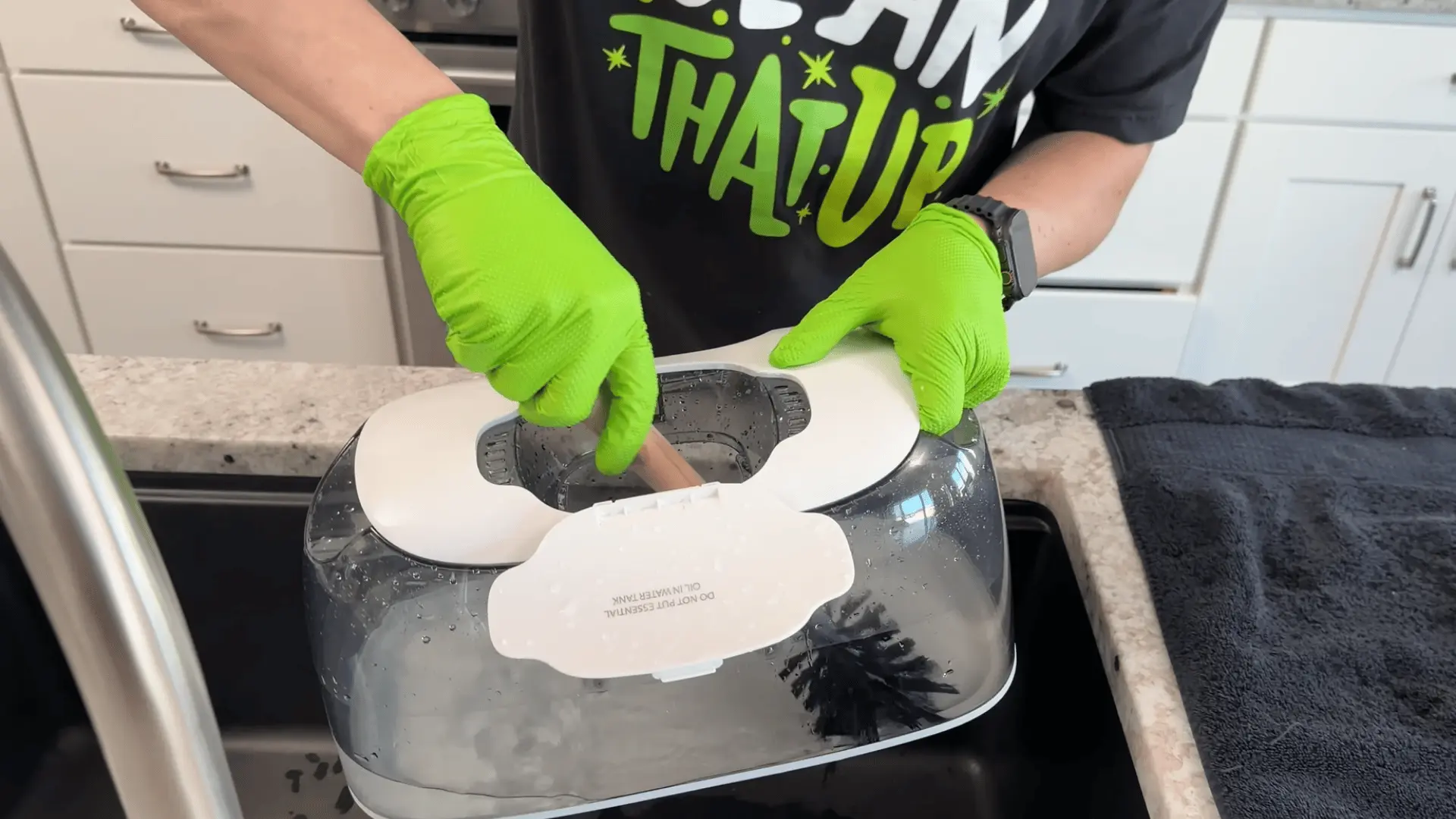 Person wearing gloves cleans a humidifier tank with a brush near a kitchen sink.