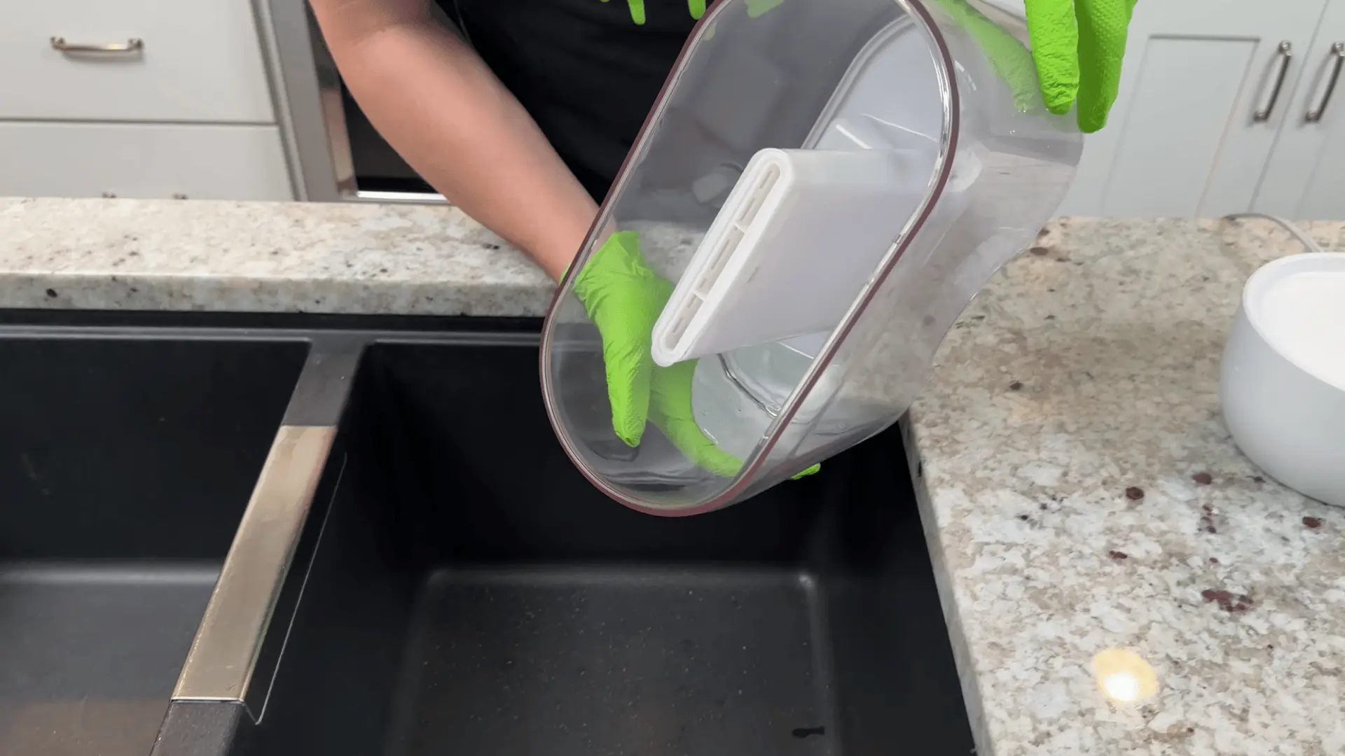 Person wearing gloves rinses a humidifier water tank over a kitchen sink during cleaning.