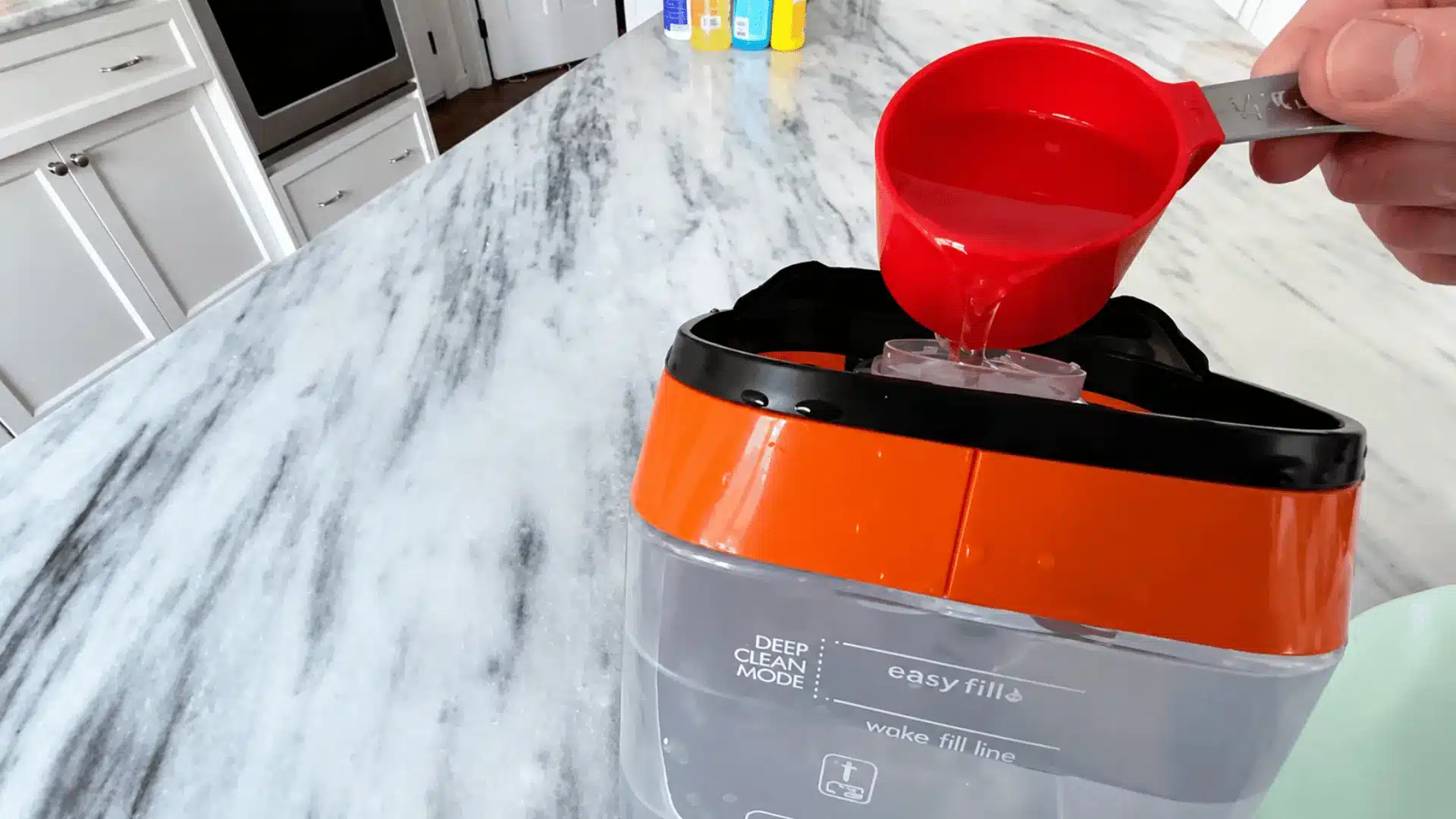 Person pouring homemade carpet cleaning solution into a carpet cleaner tank on a bright kitchen countertop before shampooing carpets.