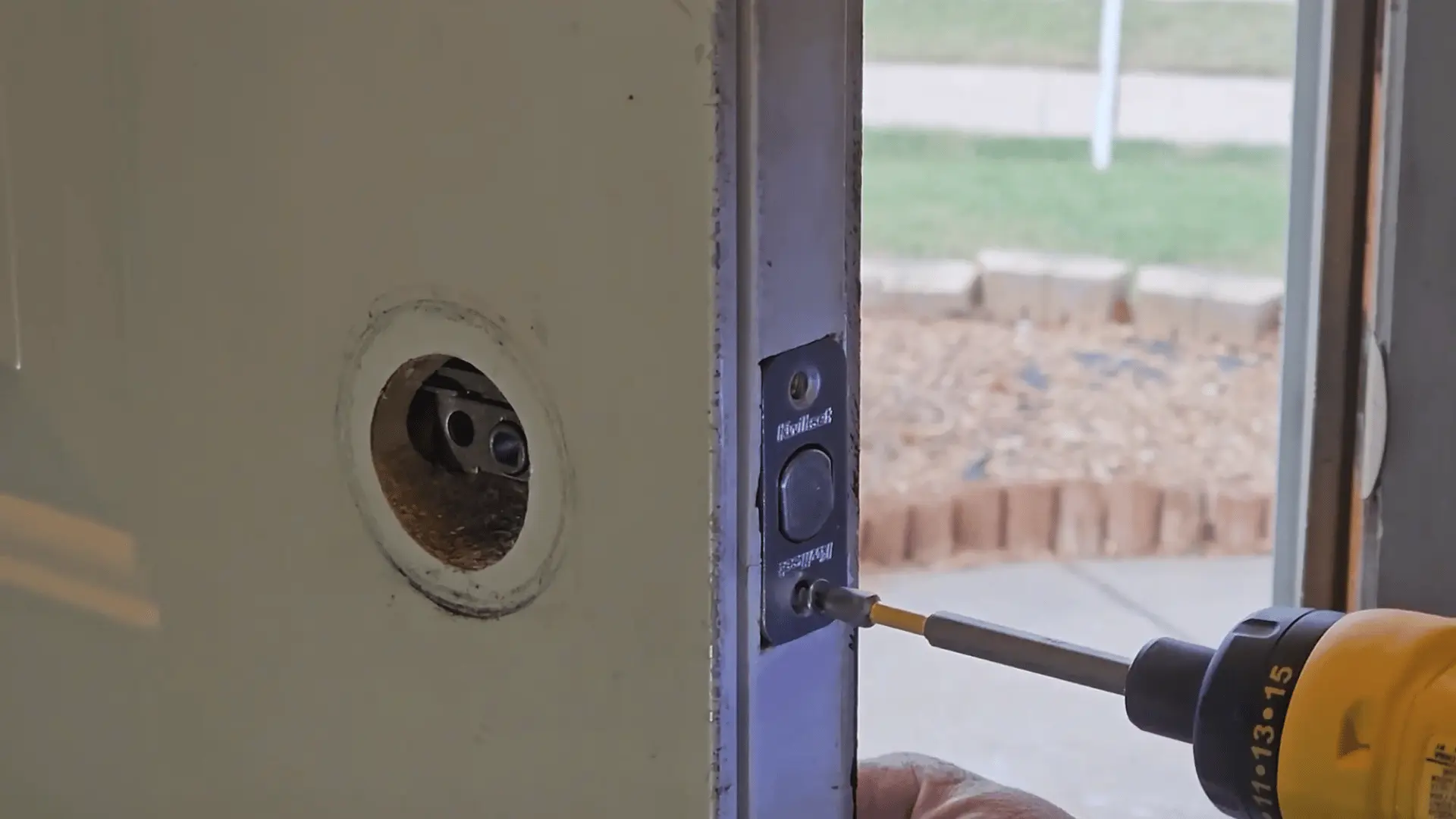 Drill tightening screws on a deadbolt strike plate installed on a door frame while aligning lock hardware through the door opening.