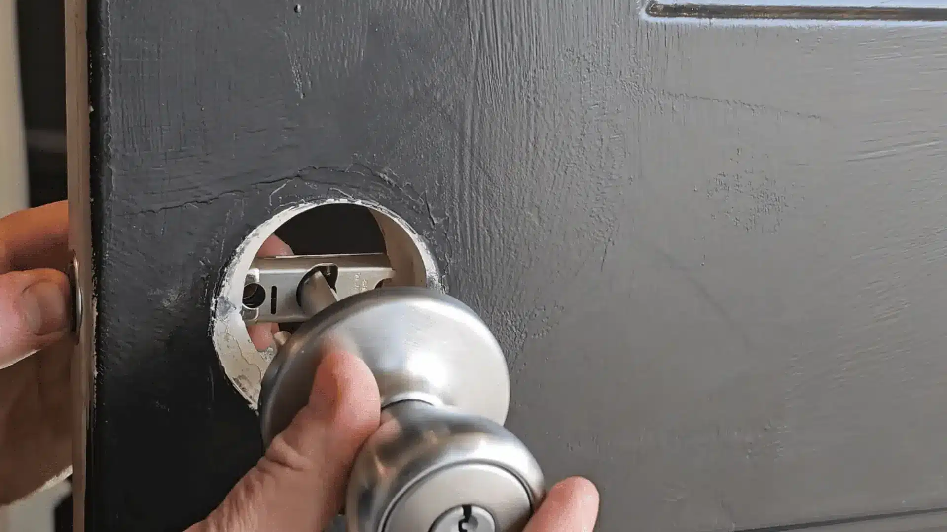Hands installing a silver door knob into a circular cutout on a grey painted door, aligning latch mechanism inside.
