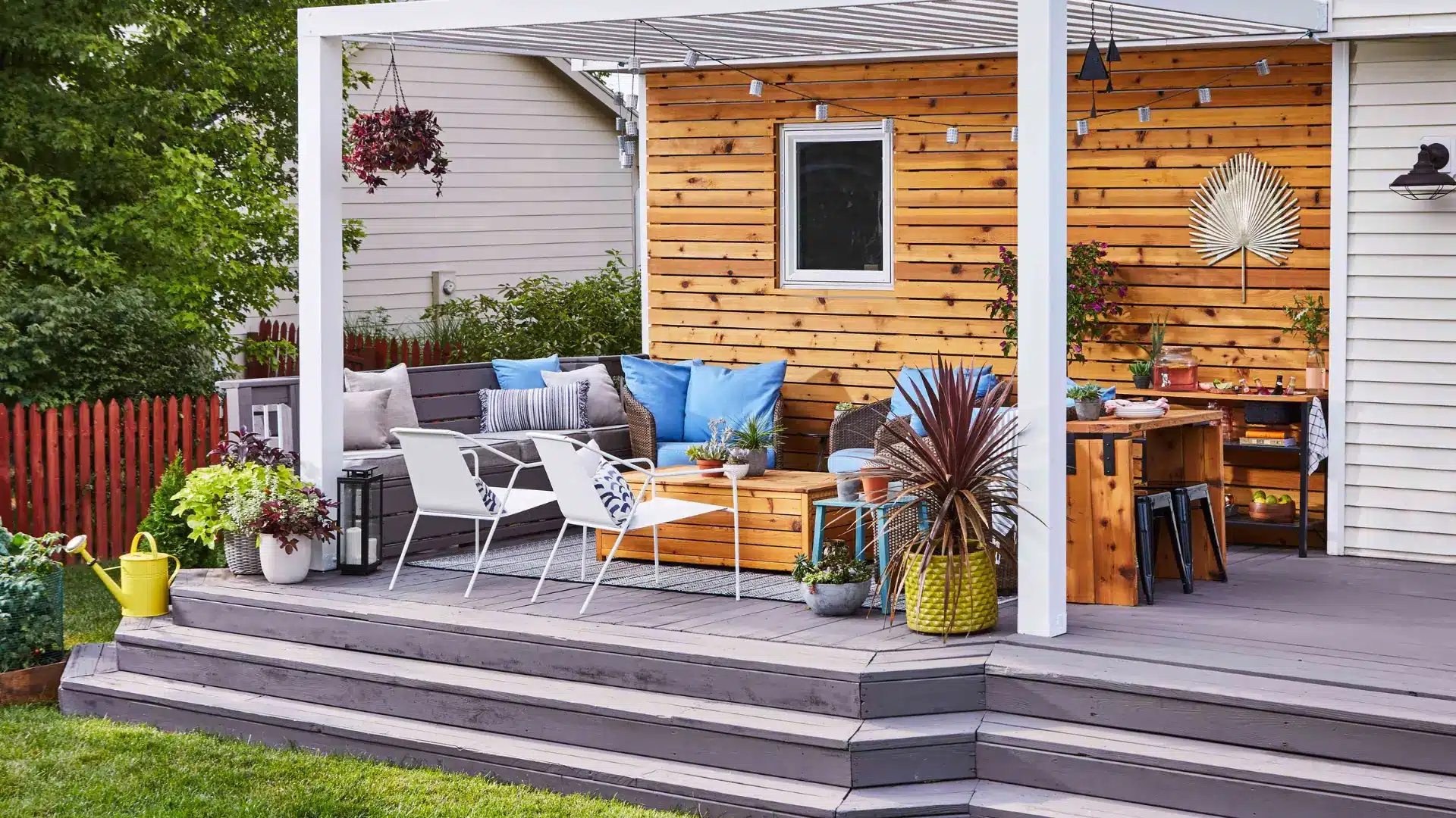 A modern wooden deck with seating, potted plants, and a white covered pergola frame.