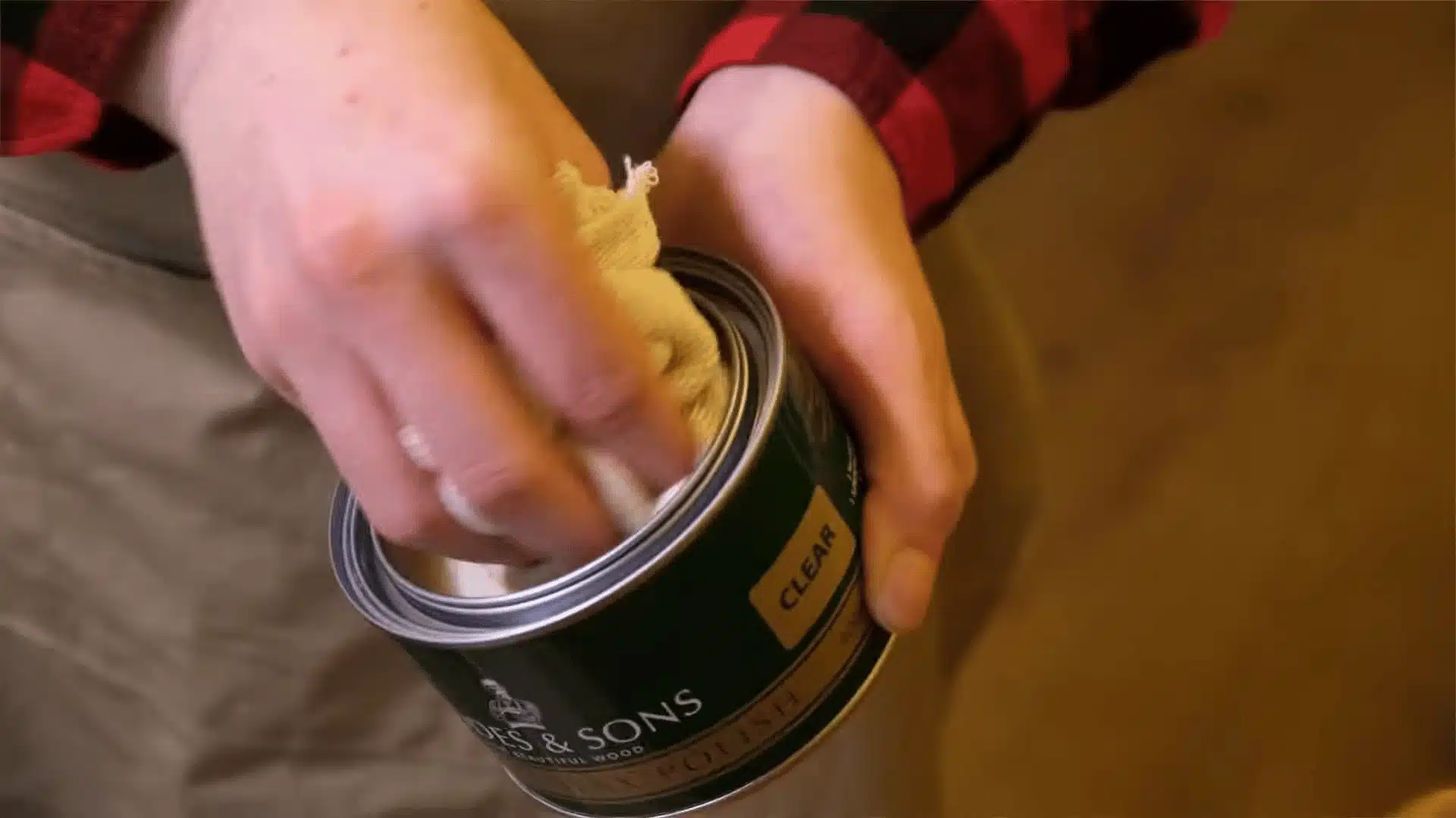 Close-up of hands wiping a cloth into an open tin of wood polish, with a green metal can labeled “clear” held in one hand.