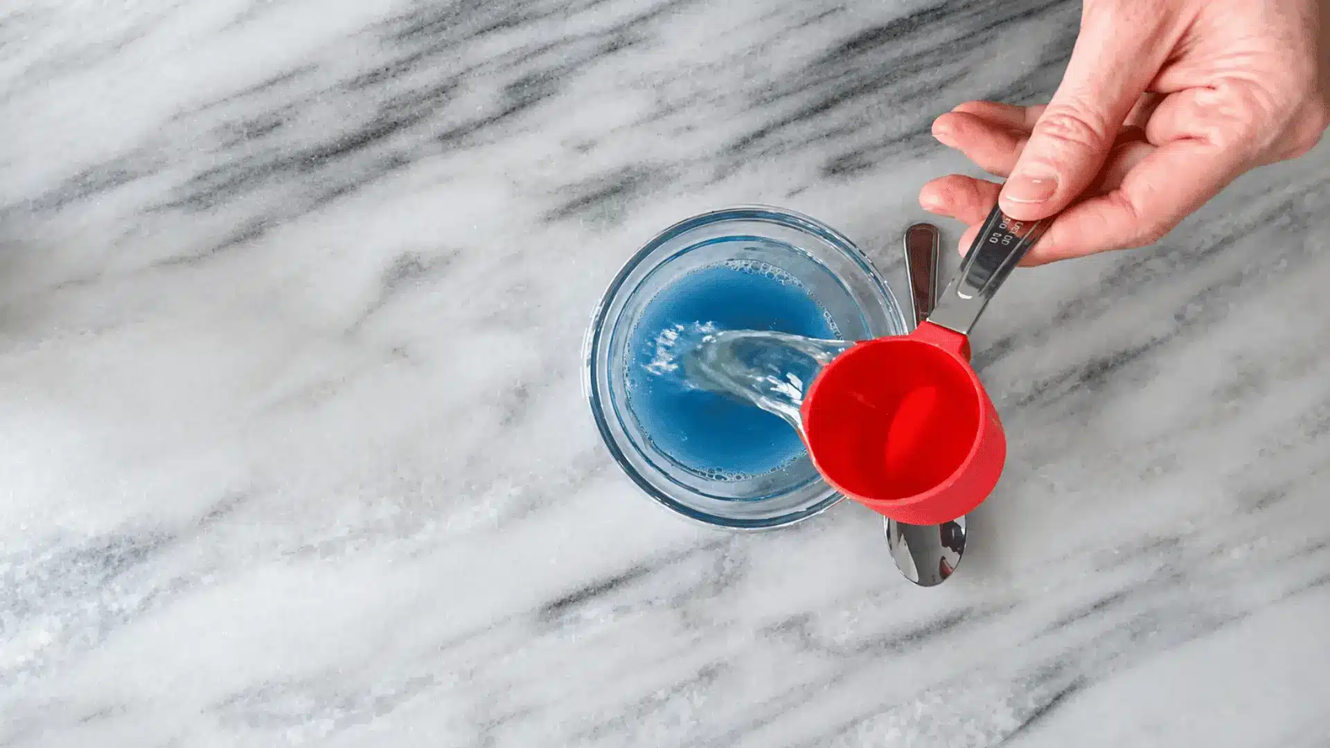Hand pouring liquid from a red measuring cup into a homemade carpet cleaning solution on a marble kitchen countertop.