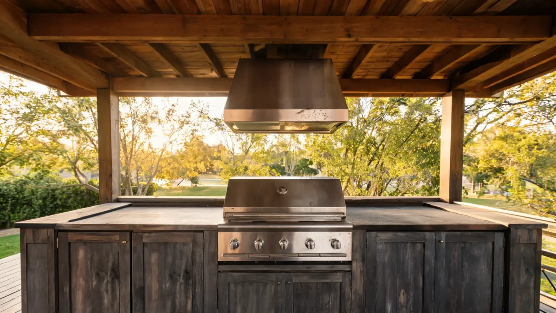 Covered outdoor kitchen setup with a stainless steel grill, vent hood, and dark wood cabinetry.