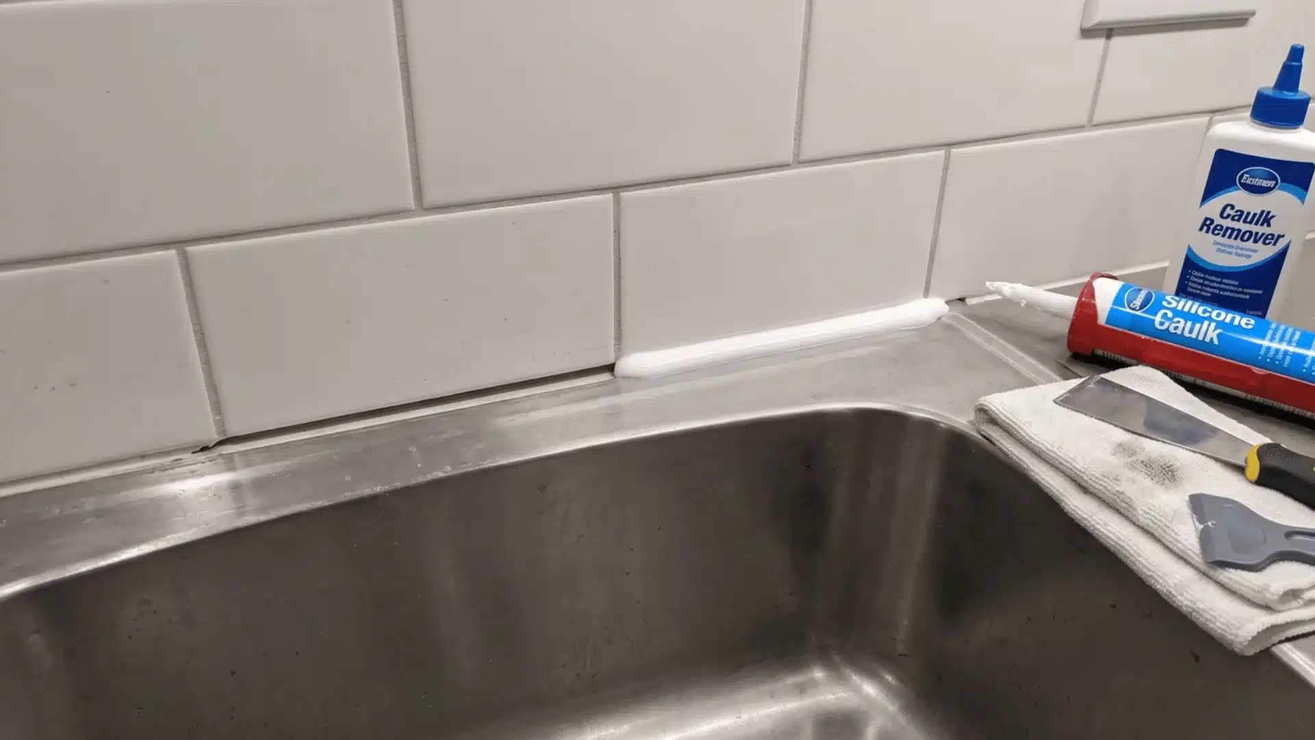 Sink joint partly recaulked with fresh silicone beside a caulk gun, scraper, remover, cloth, and finishing tool