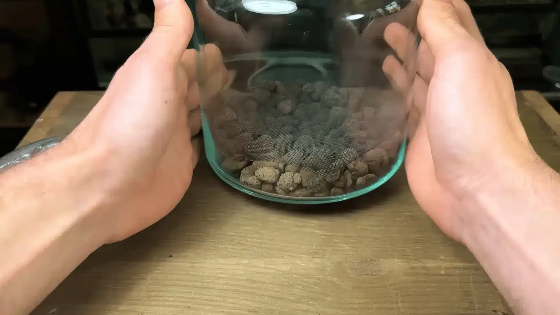 Hands holding a glass jar terrarium while placing a mesh barrier over a layer of drainage stones to separate soil from rocks.