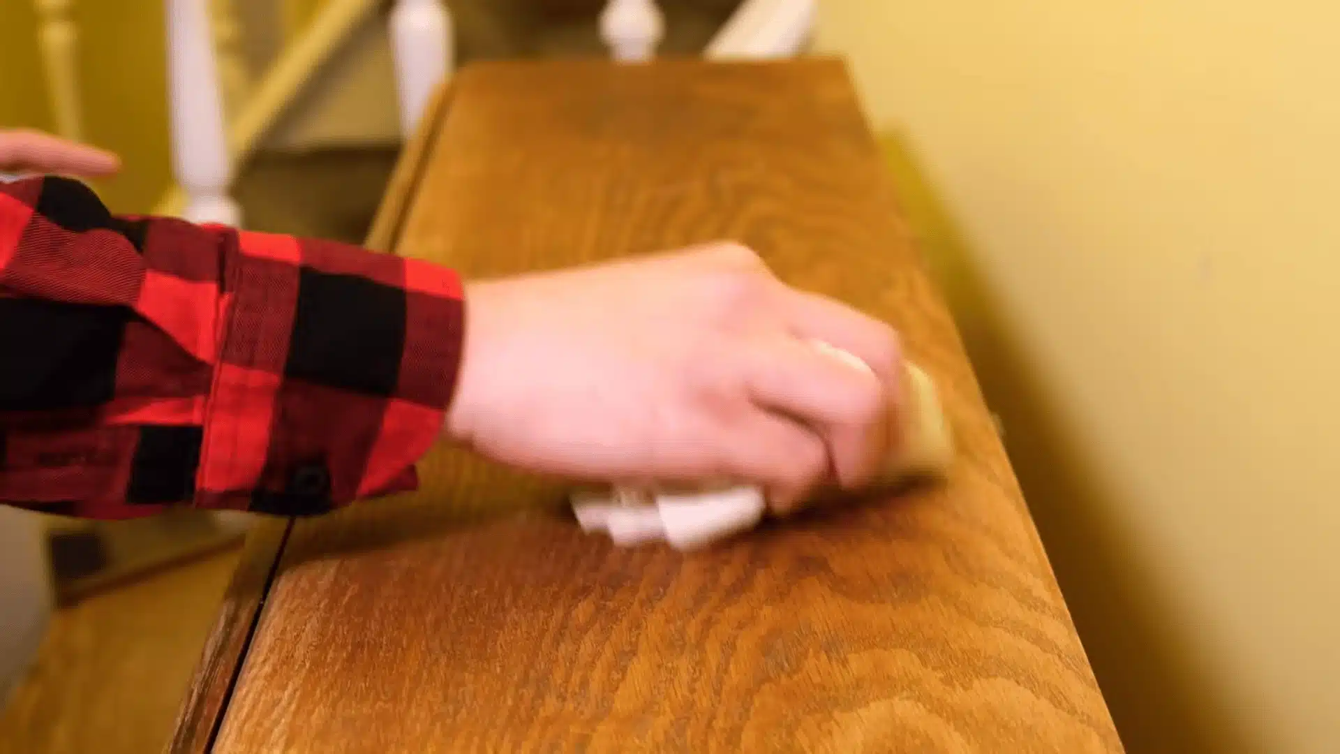 Close-up of a hand in a red plaid shirt applying polish with a cloth onto a wooden staircase banister, showing wood grain and surface shine during restoration.