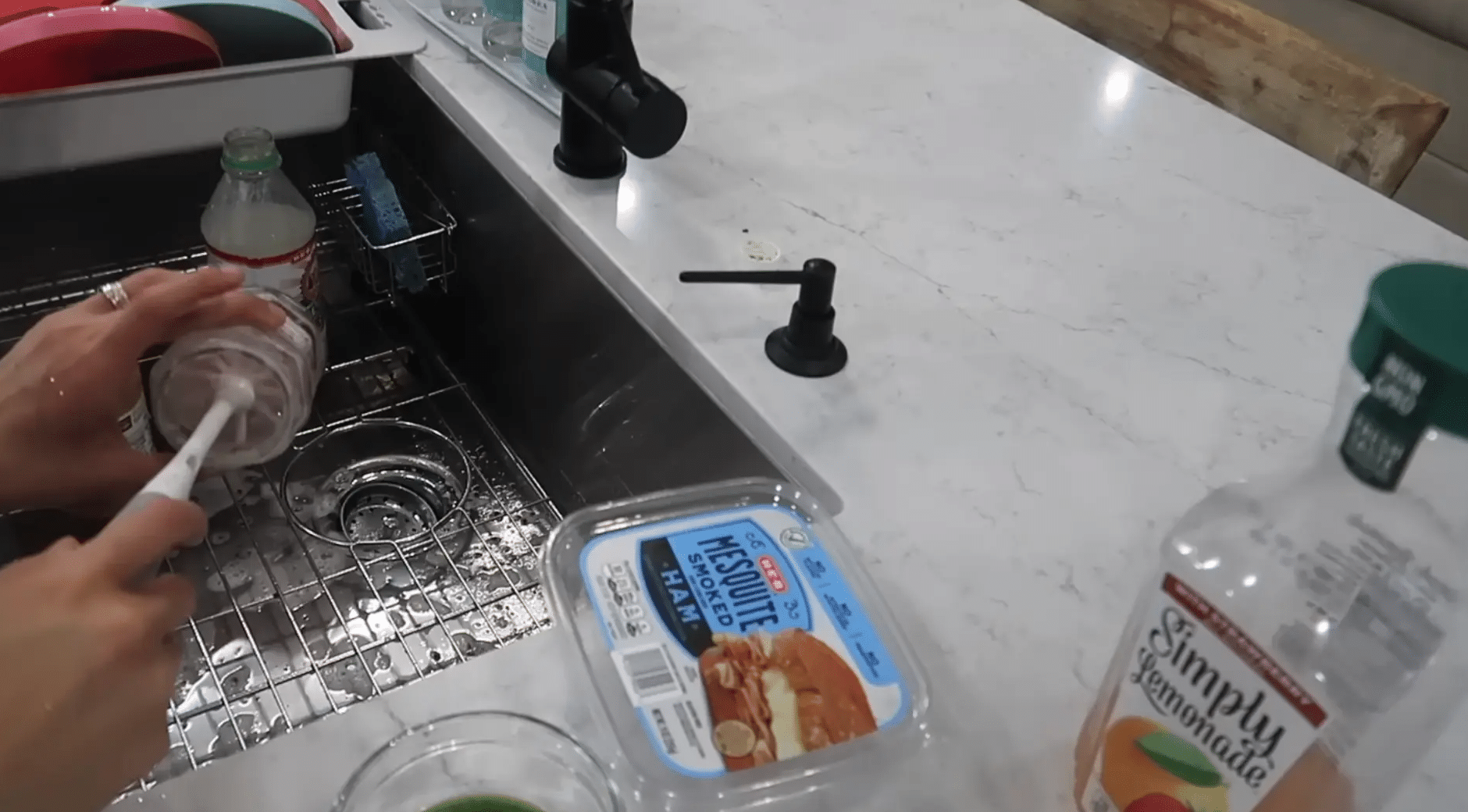 Hands washing a clear plastic container with a brush in a kitchen sink beside other labeled containers