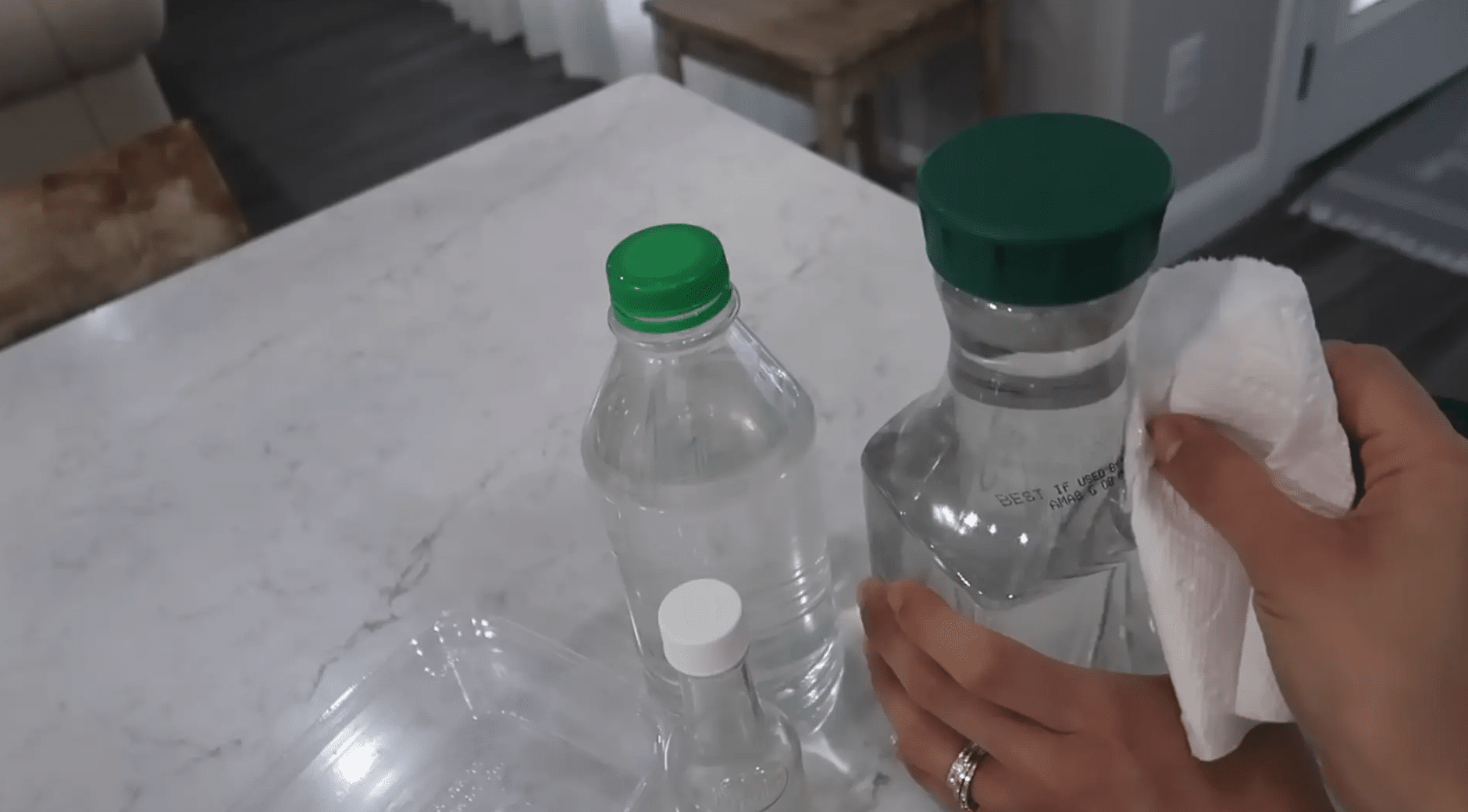 Hands wiping printed ink from a clear plastic bottle with a paper towel on a kitchen counter.